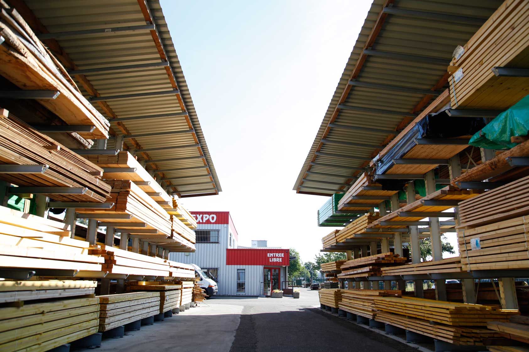 Entrepôt de bois avec des étagères chargées de planches, éclairé par un ciel ensoleillé.