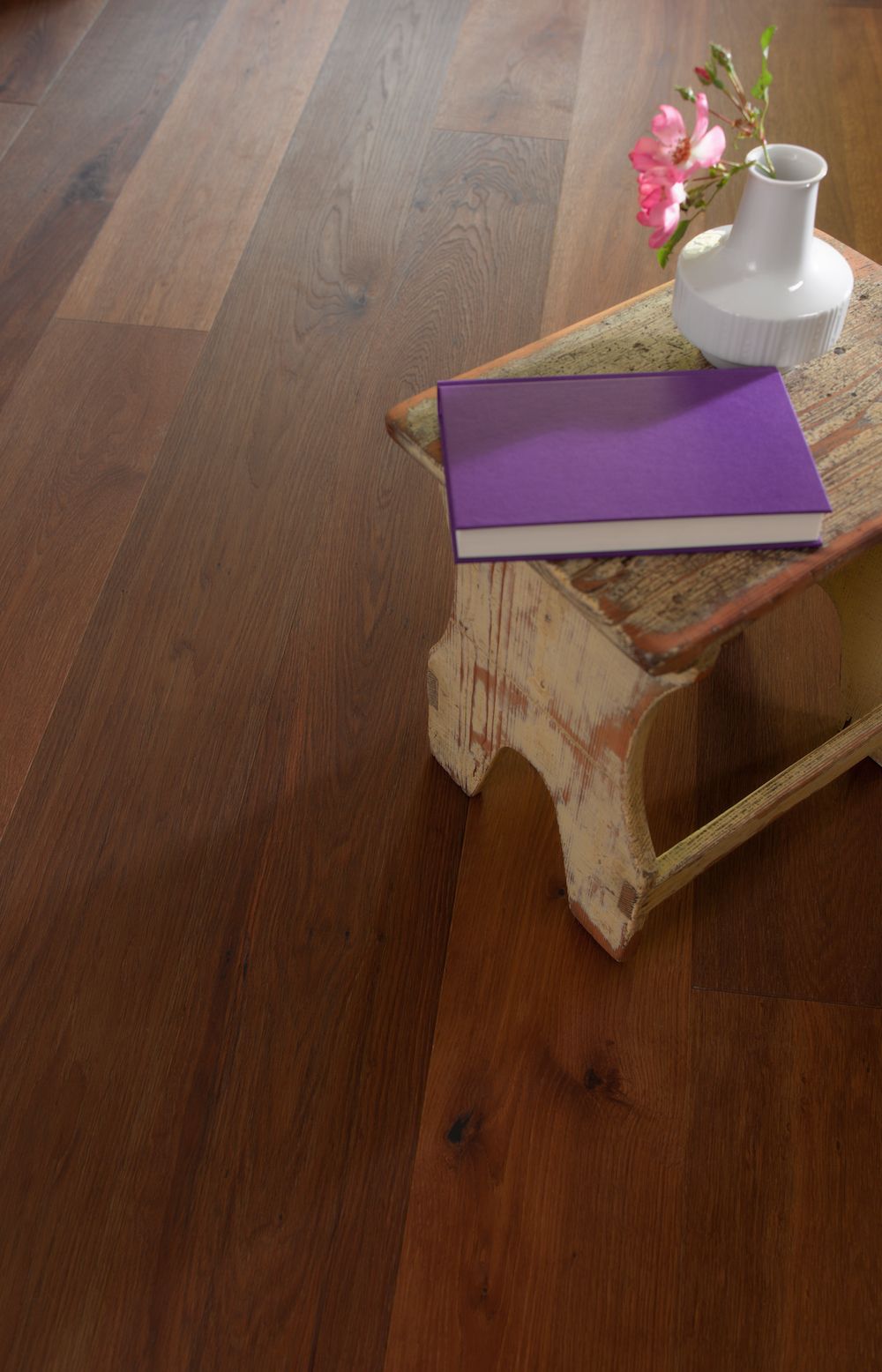 Un tabouret en bois supporte un livre violet et un vase avec une fleur sur un sol en bois.