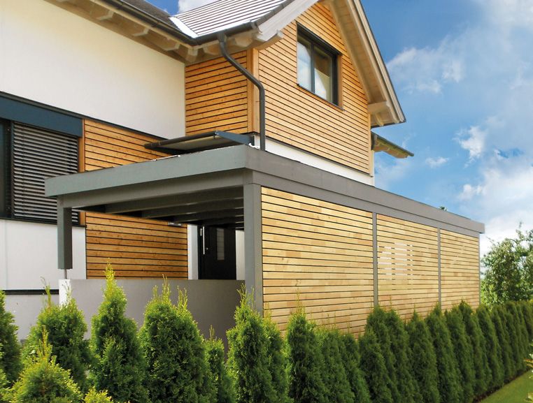 Maison moderne avec façade en bois, abri en surplomb et haie verte en avant-plan.