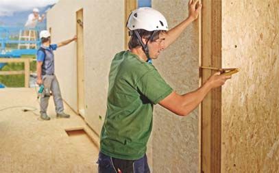 Deux ouvriers construisent un mur en bois, l'un mesure tandis que l'autre fixe des panneaux.